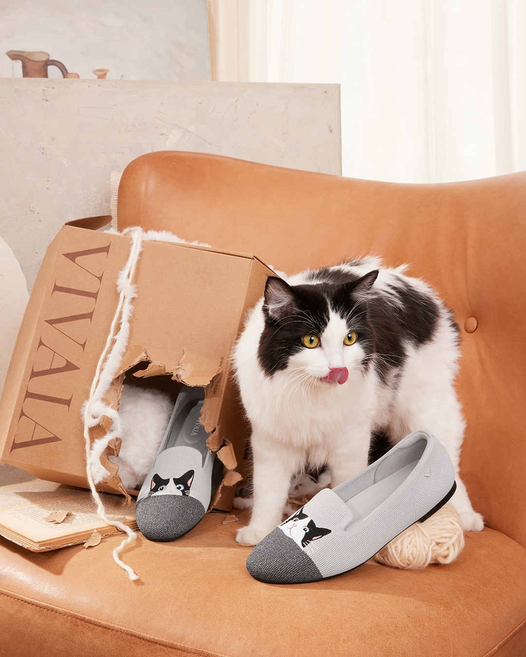 A fluffy tuxedo cat stands next to a tan leather chair beside a VIVAIA box with grey Audrey loafers featuring tuxedo cat embroidery