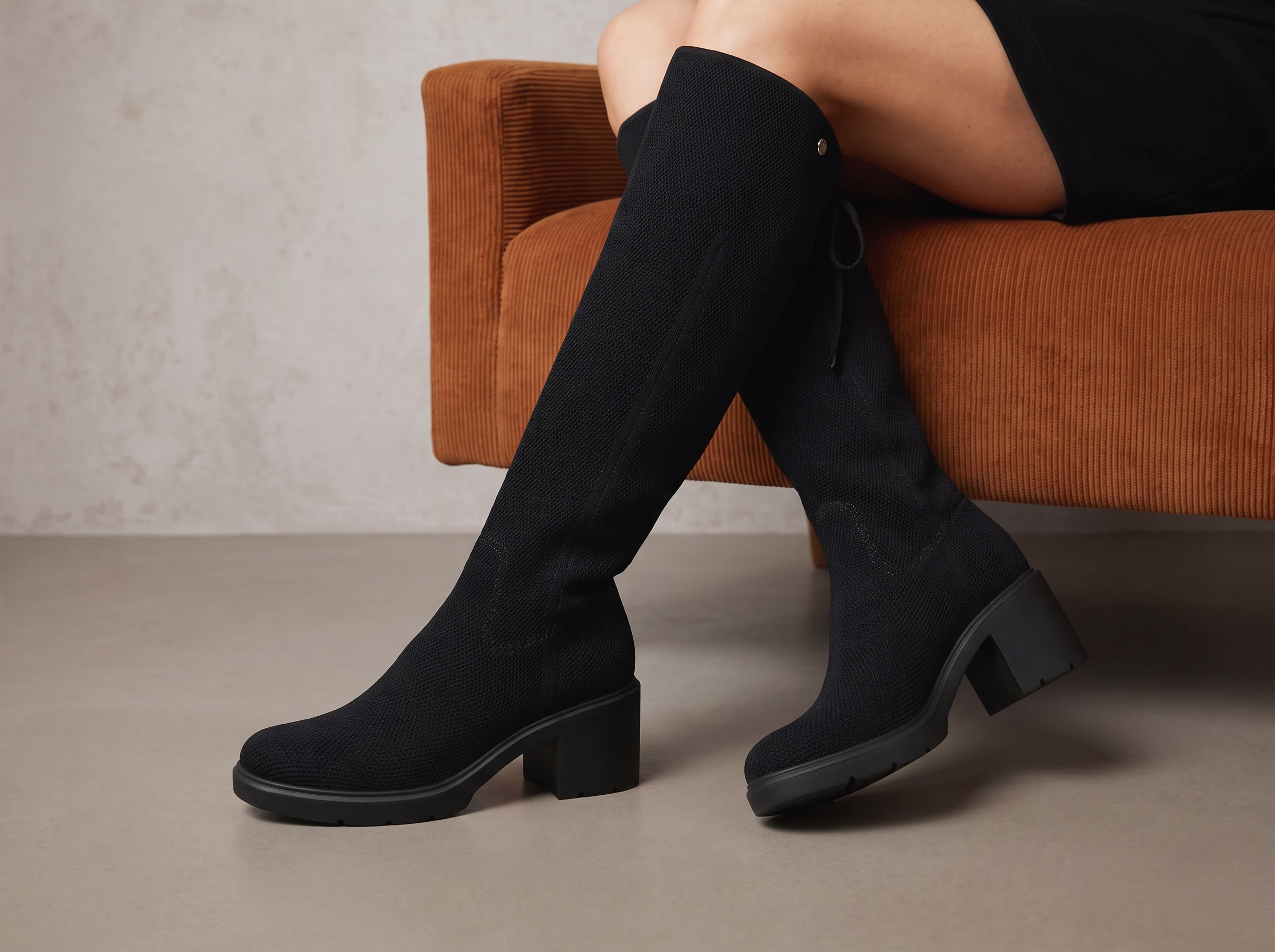 Black Gia 2.0 knit boots worn while seated beside a warm brown upholstered chair, with stable block heels fully visible