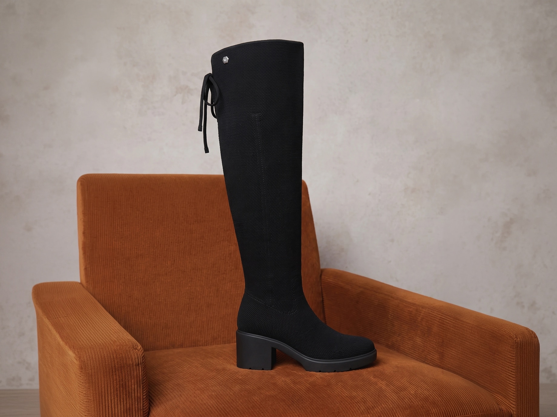 Official-style black Gia 2.0 knit boot with its shaft fully extended and top edge unfolded, on a rust-orange velvet chair