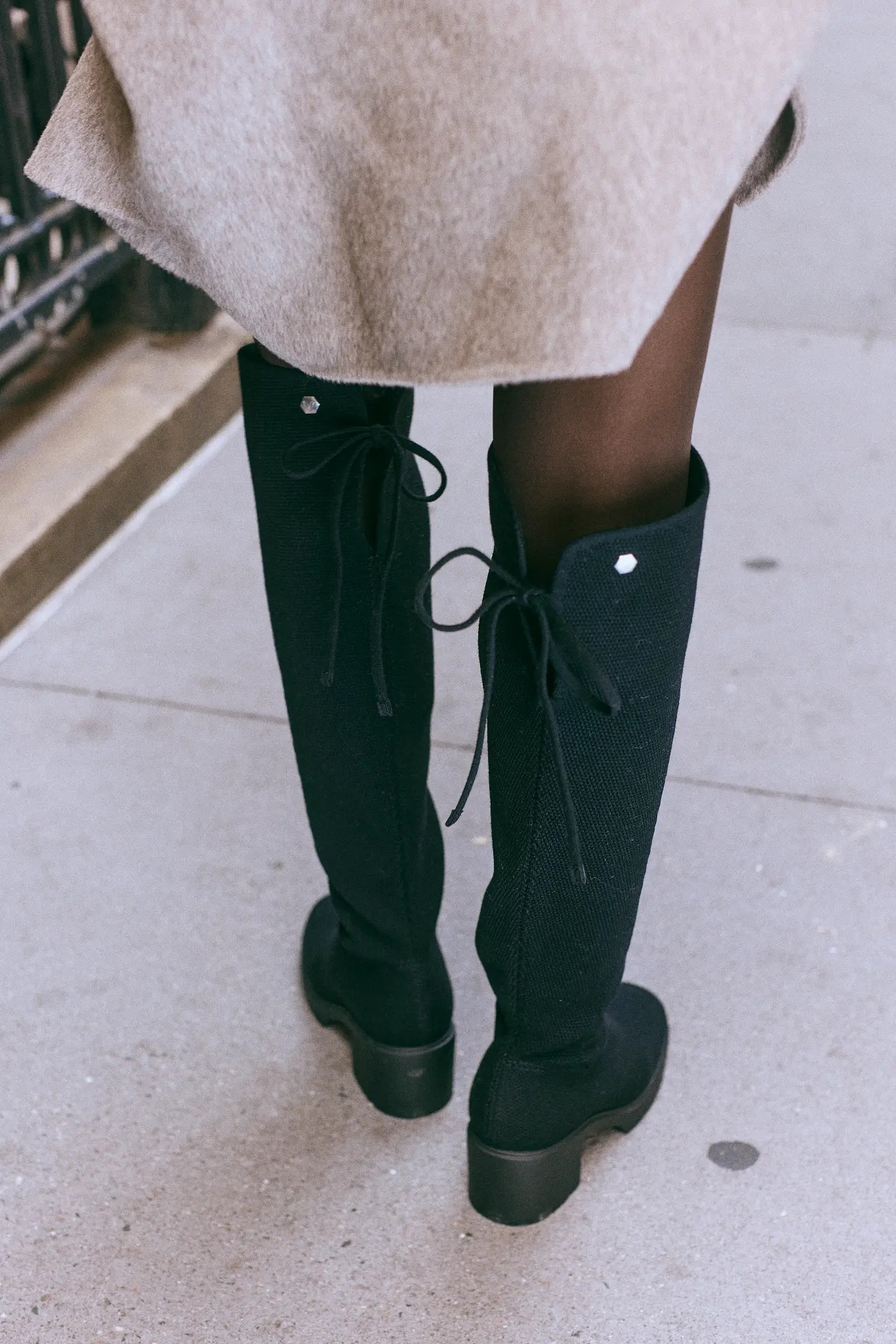 Rear view of black knit knee-high boots with bow ties, metal emblems and block heels