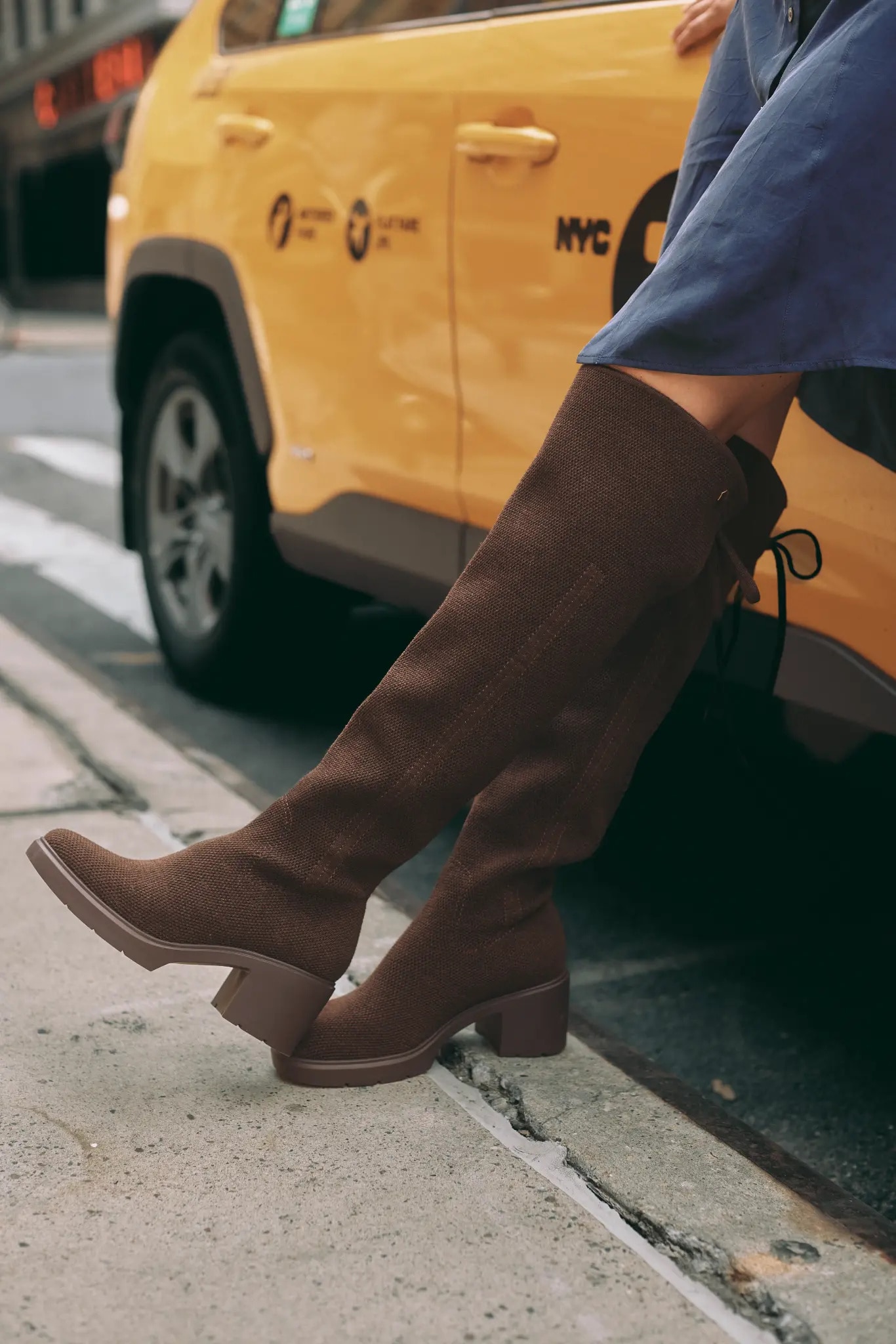 Side view of chocolate knee-high knit boots and block heels beside a New York yellow taxi